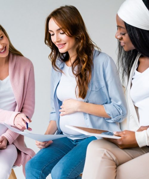 smiling pregnant multiracial women using digital tablet during prenatal class isolated on grey